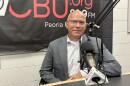 A man in a gray suit and glasses sits at a radio station desk with a microphone. The background shows a WCBU 89.9 FM banner and part of a white brick wall.