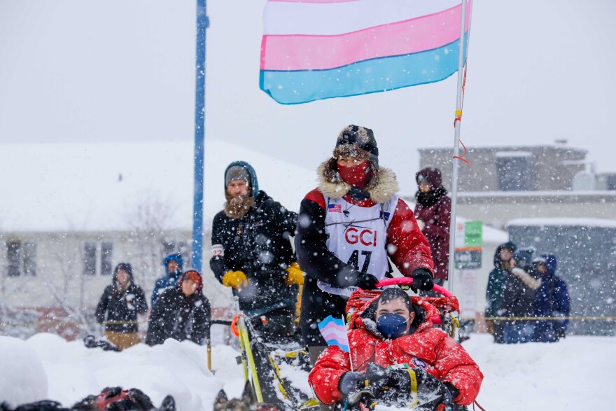 A woman in a furruffed parka drives a sled through heavy snow with a person in a thick down jacket sits in a sled in front of her