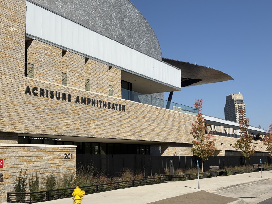 A view of the sandstone-colored brick facade of the Acrisure Amphitheater in downtown Grand Rapids. To the right of the frame, you see the roof of the amphitheater, open to the city, with a high-rise in the distance.