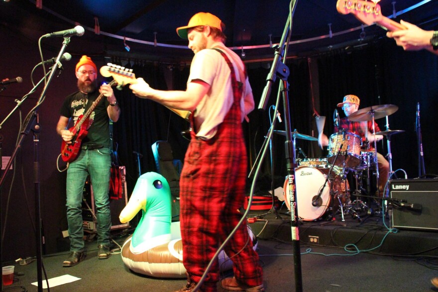 Male guitarist, inflatable mallard duck, another male guitarist wearing black and red checkered overalls and a blaze orange cap, and a drummer