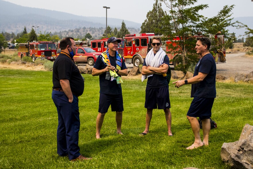 Four men are standing in a half circle on the lawn, talking to one another. Trevor Albertson is on the far-left and is wearing business casual attire. The three firefighters are in gym shorts and t-shirts. Behind them are several red firetrucks.