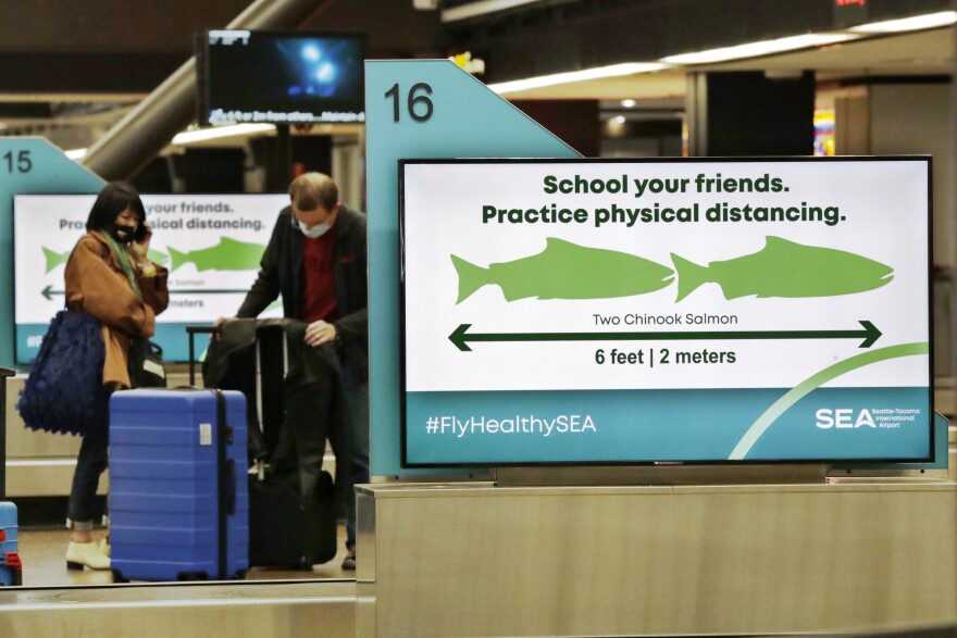 Travelers pick up luggage where signs remind them, with classic Pacific Northwest icons showing the size of two Chinook salmon, to stay six feet apart at Seattle-Tacoma International Airport Monday, May 18, 2020, in SeaTac, Wash.