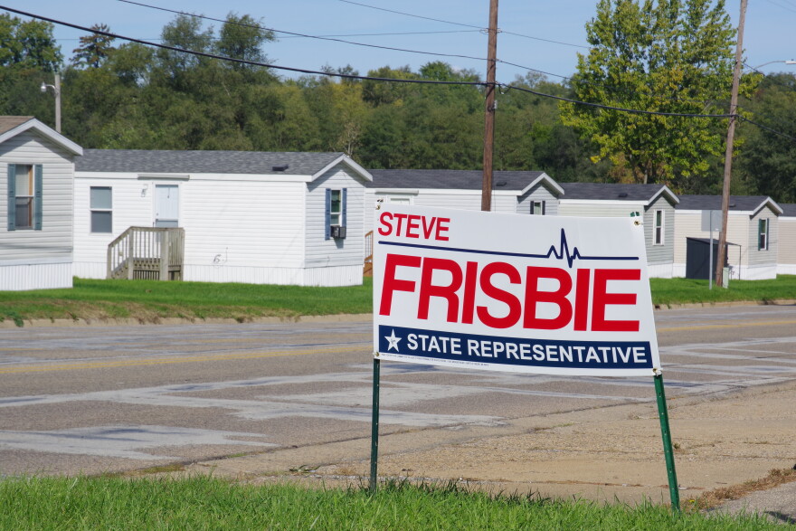 A sign for Republican State Representative Steve Frisbie of Michigan's 44th state house district stands on a green grass lawn next to a road. Behind the sign, on the other side of the road, sit multiple identical long mobile homes. 