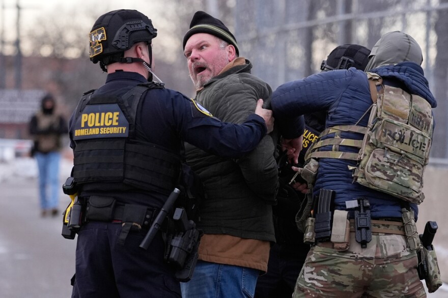 Two people in police/military uniforms detain a man in a beanie.