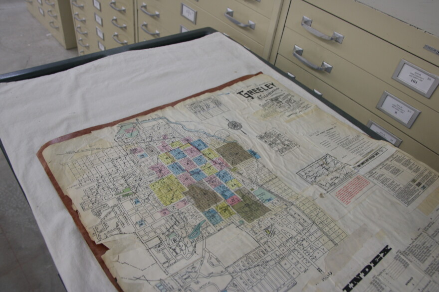A map inside a book has old, wrinkled and stained paper with several add-ons glued onto the page. The book is laying on a cart with a white sheet underneath it. Next to it are some filing cabinets. 