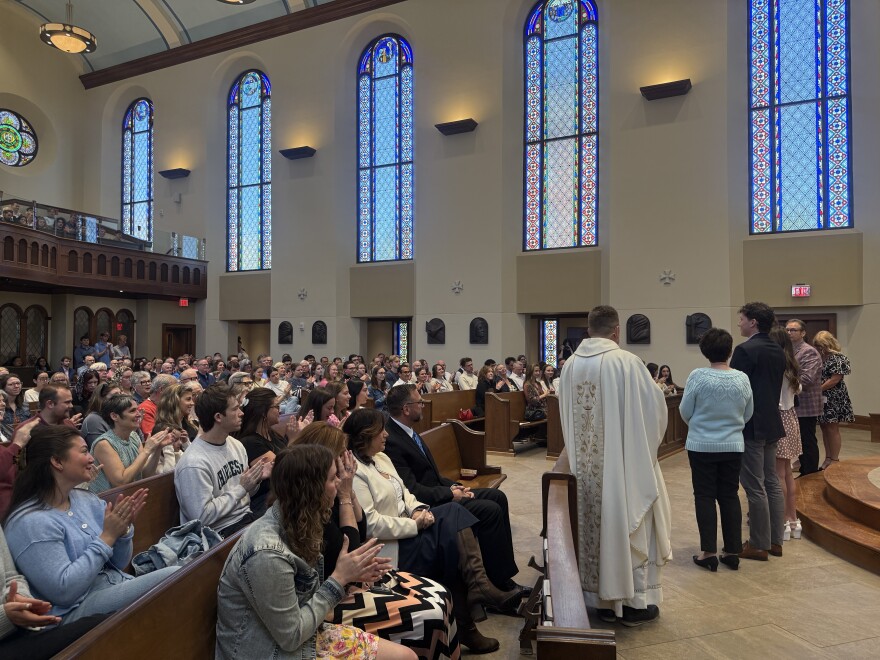 A crowd of people fill a church as a priest in white robes and a handful of people stand at the front.