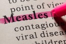 illustration represents close up of a book page with the words Measles, contagious and viral disease, with a red highlighter marking through the word Measles. 