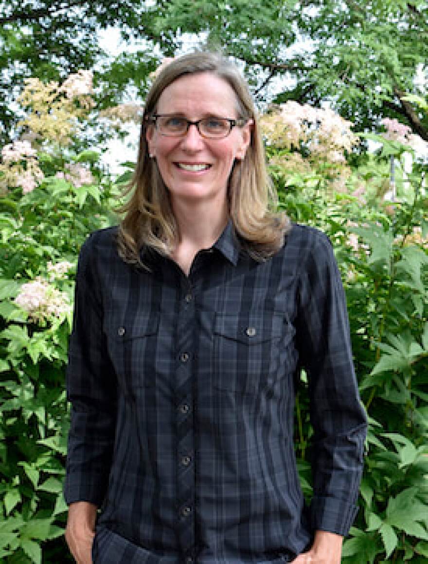 Heather Holm with a blue shirt and glasses stands in front of a row of native flowering plants.