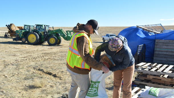 One person hold open a large white bag as another pours seeds from a paper bag into it.