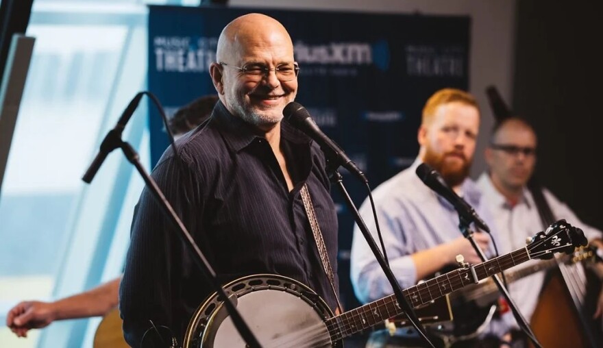  Sammy Shelor standing in front of a microphone with a banjo hanging from his shoulder. 