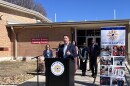 A man in a navy suit jacket speaks at a podium with the North East ISD emblem. Four women and a man in business attire stand behind him. Behind them is a school building with a banner that reads "Wilshire Safety Training Center."