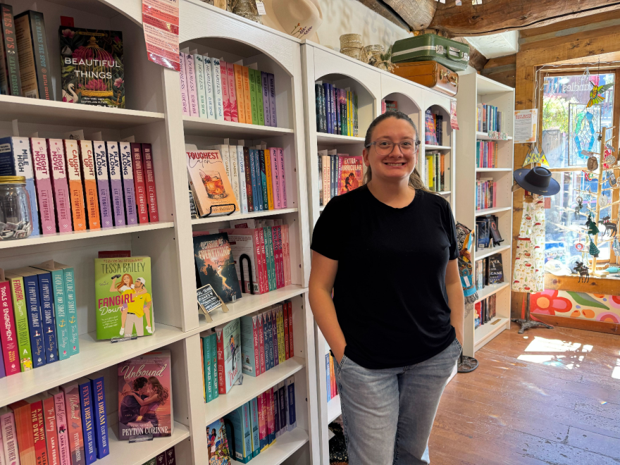 A woman stands in front of a row of neatly-organized bookshelves inside a store. The visible bookshelves are full of brightly-colored romance novels.