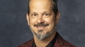 A smiling man with a dark red checkered jacket and a black shirt looks toward the camera.