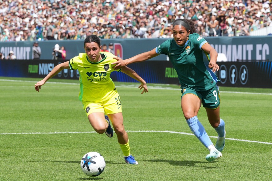 Two women in soccer uniforms chase after a ball.