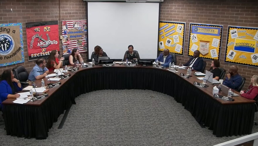A group of people sit around a large U-shaped table in a meeting room, with colorful bulletin boards and a blank projector screen on the back wall.
