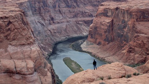 A man stands on a red rock cliff looking out over a blue river below