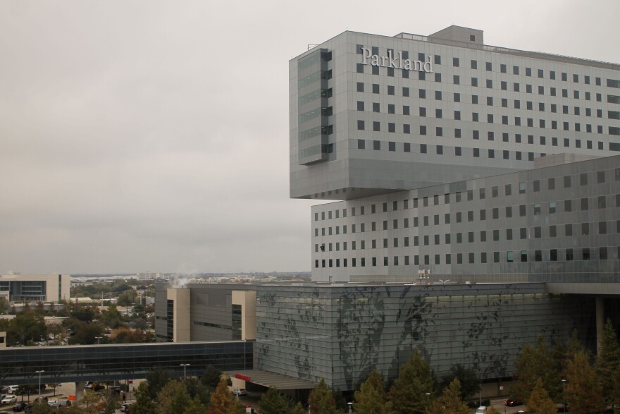 The Parkland Hospital building on an overcast day. The building includes a large portion that's primarily glass window panes with a gray decal of plant silhouettes covering them. One top of that are two gray rectangular shaped structures that are sitting perpendicular to each other with dozens of small windows on them. The top gray structure has a sign that reads "Parkland" in the upper left corner. Trees and other building surround the hospital.