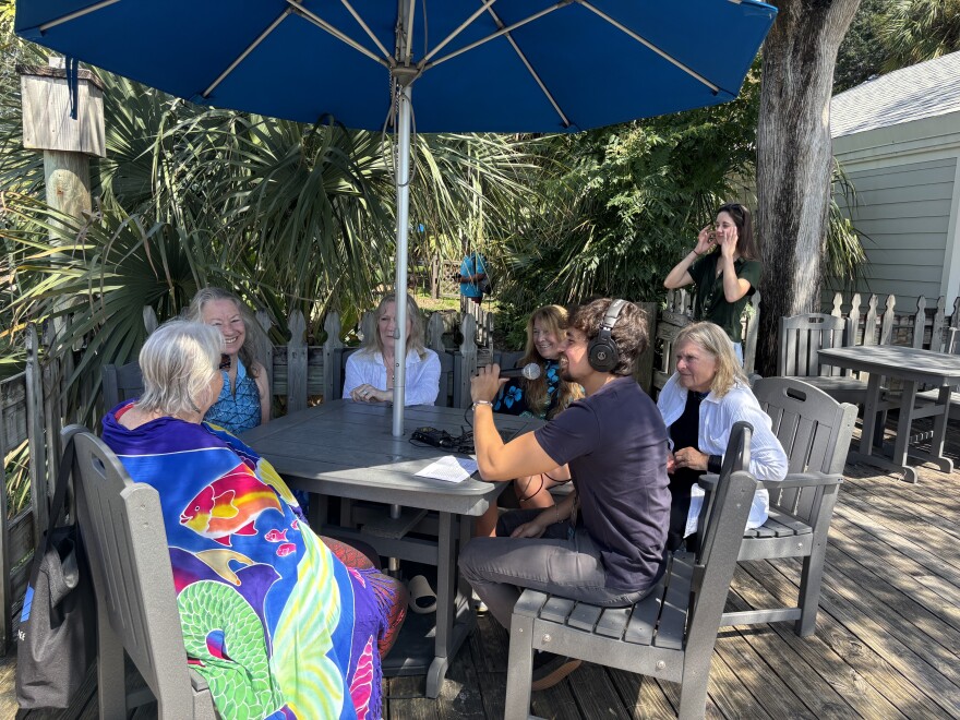 Sky Lebron sits around a table talking to the five mermaids under an umbrella. The women have wet air from swimming in the spring just a bit earlier.