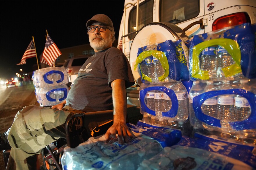 Rob McFadden sits outside East Palestine High School as community members line up to attend a meeting about the train derailment. He brought packages of bottled water to give away.