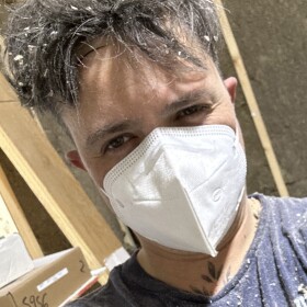 Person covered in sawdust wearing a ventilation mask looking into the came with bemused expression