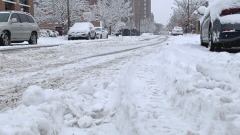 A close-up low shot of a snowy downtown Columbia street. Tires tracks are visible in the snow. Cars parked along each side of the street are covered in snow. 