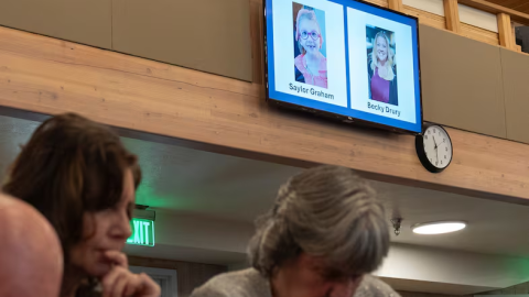 Two people bow their heads. On the wall behind them images of Saylor Graham, 7, and Becky Drury are displayed on a screen.