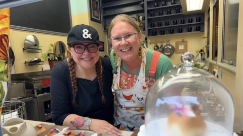 Alicia Dawn, wearing a black shirt and cap, and Erica Idso-Weisz, wearing a colorful apron, smile together behind the counter at Happy Luncheonette in Medford. Small plated appetizers sit in front of them, with a covered pastry stand in the foreground and the restaurant kitchen in the background.