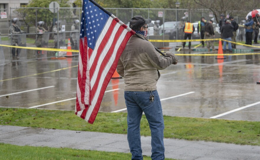 A protestor holding a U.S. flag stands outside the WA state capitol in Olympia while National Guard and Washington State Patrol guard the fence during the first day of the legislative session on Jan. 11, 2021. 