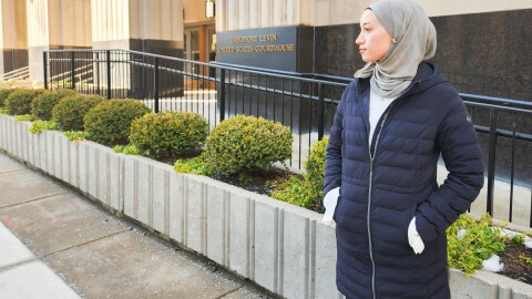 A young woman in a gray hijab and navy coat stands in front of the Theodore Levin United States Courthouse in Detroit. Behind her is a railing and decorative shrugs. She is looking to the left of frame. 