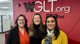 Three women stand shoulder to shoulder smiling at the camera. A large red wall sits behind them in an office. 