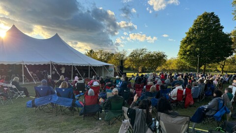 A group of people in lawn chairs watch an orchestra perform under a tent outdoors
