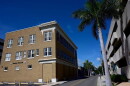 A tan, three-story building stands on the left, next to a road lined with palm trees, a parking garage on the other side of the street.
