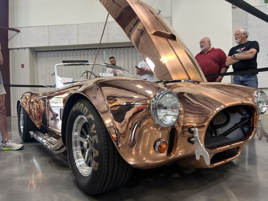 A shiny, copper, antique car sits indoors with four men to the right looking at it. 