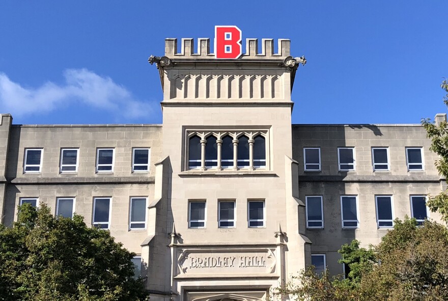 A stone academic building with a “B” in red on top, reading “Bradley Hall” above the entrance, with trees and a clear blue sky.