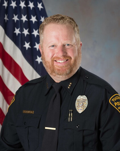 A portrait of Chris Dennison in his police uniform is shown, with an American flag behind him and a gray background.