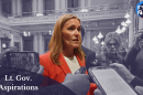 A photo of Winnie Brinks in front of a monochromatic background. In the bottom left corner is a semitransparent semi-circle with the words "Lt. Gov. Aspirations." In the top right corner is the It's Just Politics logo.