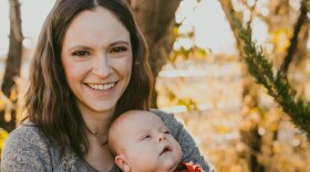 A woman and her baby are pictured in front a blurred forest scene near sunset. She's smiling toward the camera. She has shoulder-length brown hair that's parted in the center and is wearing a grey sweater. Her baby looks toward the right and has chubby cheeks and is wearing a red and black plaid long-sleeve top and blue jeans.