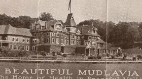 An archival publicity photo showing a large, multi-story Victorian-style hotel building with text at the bottom that reads 'BEAUTIFUL MUDLAVIA, The Home of Health in Peaceful Valley, KRAMER, INDIANA.'