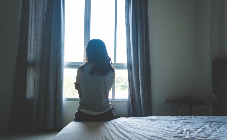 A woman with long, dark hair sits on a bed, looking out the window. Her back is to the camera.