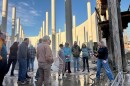 A woman speaks to a group of people standing inside a massive, roofless round structure filled with columns. 