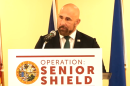 bald man with trimmed full beard in a suit speaking behind podium with a sign that reads operation senior shield