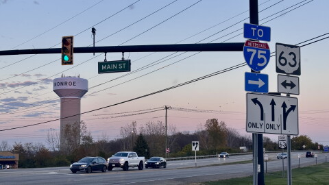 Cars sit stopped at a red light with a sunset and Monroe water tower in the background.