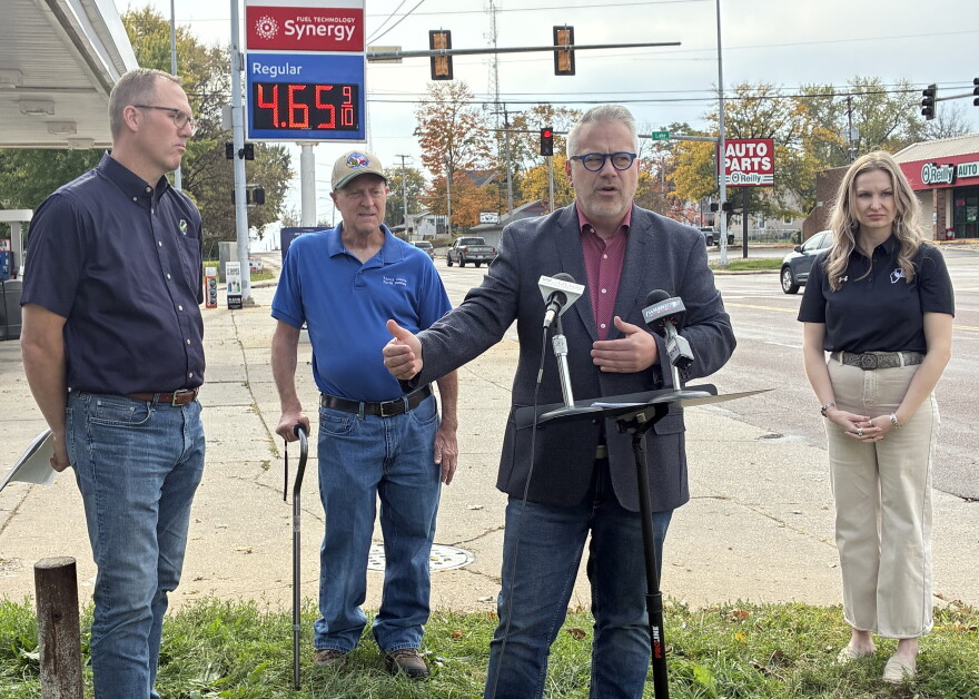 A man in a blazer speaks at a podium with two microphones, flanked by a man in a blue polo, an older man with a cane, and a woman in a black polo, with a gas station price sign in the background.