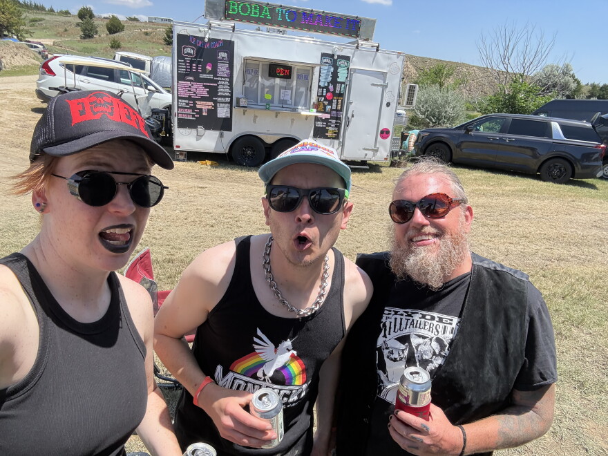 Three people stand grouped together wearing black clothing. One wears black lipstick and another wears a chain-link necklace. They stand in front of a food truck outside in a dry field.