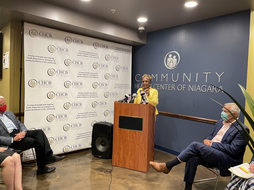 Dr. LaVonne Ansari stands at a podium wearing a bright yellow jacket. Behind her are logos for the community health center and in the foreground other individuals are seated.