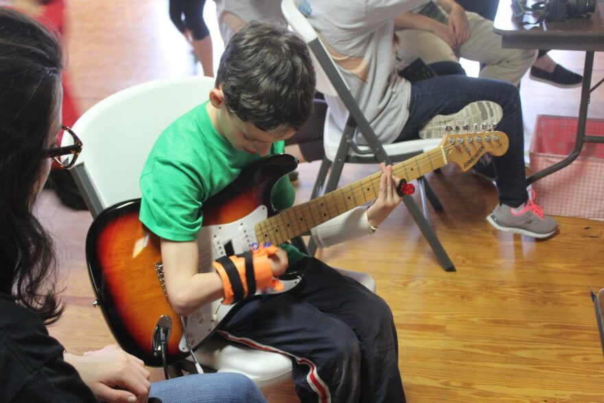 Jonathon Podber, 10, plays the electric guitar using one of the prosthetics created by UF GRiP. Because of his upper-limb difference, he doesn't have fingers on his right hand to hold a guitar pick.The orange device has a pick attached so can strum pain-free. (Carly Breit/WUFT News)