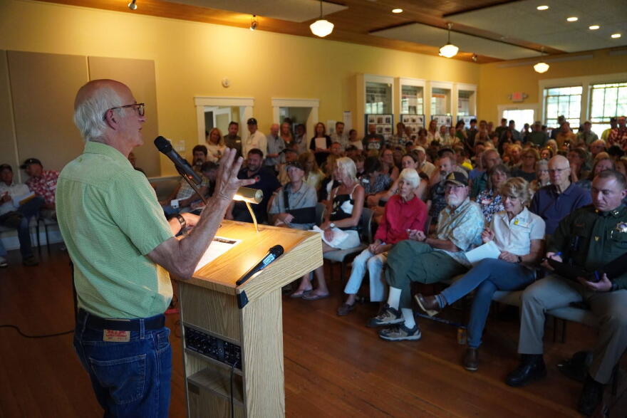 A man stands at a podium, speaking into a microphone in front of a full room of people, some of whom are standing lined around the walls.