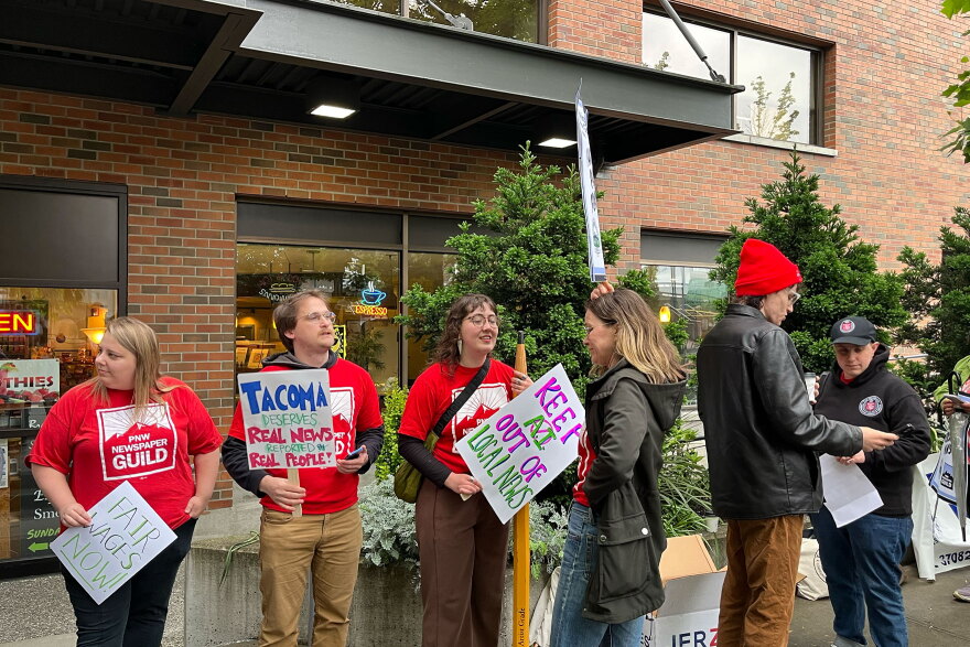 A group of people in red shirts stand in a line. They are holding signs that say things like "Keep AI out of local news."
