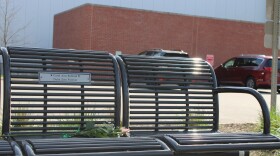 A photo is taken from low to the ground of one half of a memorial bench. There is a flower laid on the center seat and the plaque reads, "Carol Ann Rofstad Delta Zeta Forever."