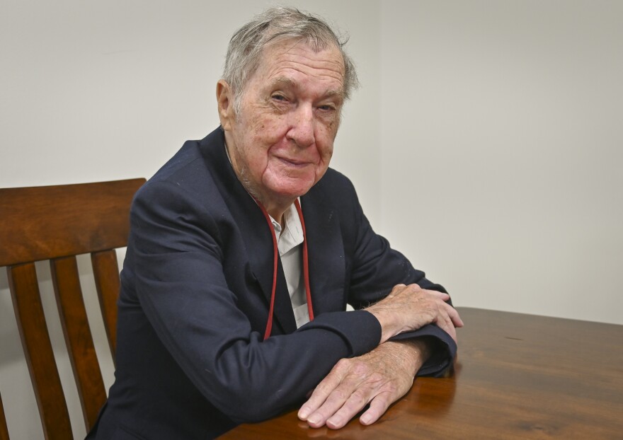 A man wearing a blue suit coat sits at a table with his arms folded on it. He is smiling and looking at the camera.